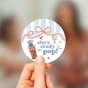 以下が含まれることがあります： 水色と白のストライプの背景に、青いリボンが付いた茶色のコカ・コーラのボトルが描かれた丸いステッカー。「she's ready to pop!」というテキストは青と赤で書かれています。赤いハートと星がステッカーを飾っています。