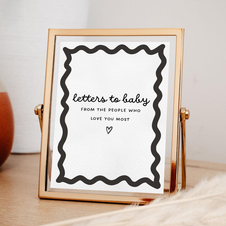 May include: A gold-coloured picture frame with a white insert. The insert has a black wavy border and text that reads "letters to baby FROM THE PEOPLE WHO LOVE YOU MOST" with a heart symbol. The frame is on a wooden surface.