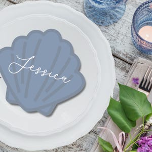 May include: A light blue seashell-shaped place card with the name "Jessica" in white script sits on a white plate. The plate is part of a table setting with a candle, silverware, and floral accents. The overall theme is a coastal or beach-themed event.
