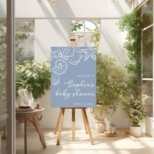 May include: A baby shower sign on a wooden easel. The sign is light blue with white seashell illustrations and text that reads "Welcome to Sophia's baby shower, April 12, 2030."