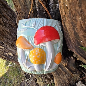 May include: A ceramic hanging planter shaped like a mushroom with a light blue background and a red, orange, and yellow mushroom design. The planter has a brown leather strap for hanging.