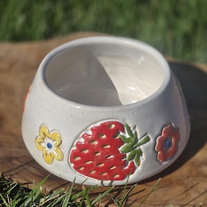 May include: A handmade ceramic bowl with a white exterior. The bowl features a red strawberry design with green leaves and yellow and red flower accents. The bowl is sitting on a wooden surface.