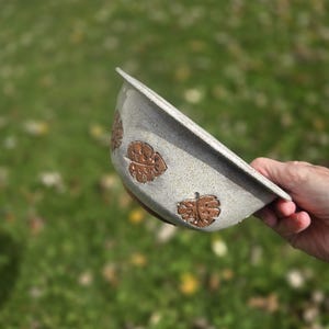 May include: A speckled gray ceramic bowl with a brown interior and leaf-shaped cutouts. The bowl is held at an angle, showcasing the design against a blurred green background.