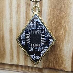 May include: A gold chain necklace with a diamond-shaped pendant featuring a black circuit board inside. The circuit board has a small square chip with the text "74HC138" printed on it.