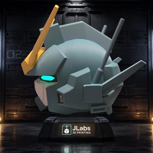 May include: A light blue robotic head sculpture featuring a prominent yellow V-fin antenna, glowing cyan eyes, and angular mechanical details. The head rests on a black pedestal base with a label that reads JLabs 3D PRINTING in white text.
