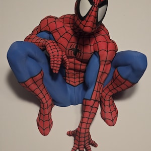 Op de afbeelding: Een Spider-Man-figuur in een gehurkte houding, met een rood en blauw pak met een zwart webpatroon en een zwart spinnenembleem op de borst. Het personage heeft grote witte ogen en is afgebeeld in een dynamische, laag bij de grond positie.