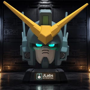 May include: A blue and yellow robotic head bust featuring a V-shaped crest, glowing cyan eyes, and a dark gray base with a label that reads JLabs 3D PRINTING. The design includes geometric panels and antenna-like structures on the sides of the head.