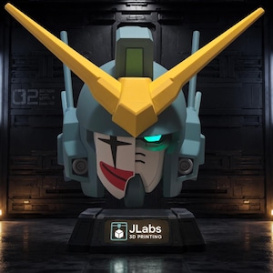 May include: A stylized robot head bust featuring a blue helmet with a large yellow V-shaped crest. The face is split, with one side showing a white clown mask with a red smile and black eye markings, while the other side has a glowing cyan eye. The base displays the text JLabs 3D PRINTING.