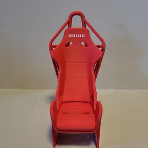 Puede incluir: Asiento de carreras rojo con un marco de metal negro y la palabra "BRIDE" impresa en el reposacabezas.