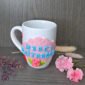 Peut inclure: Tasse en céramique blanche ornée d'une bande décorative rose avec le texte "MERCI MAITRESSE" en lettres bleues. La bande comporte des cœurs et une rose rose. Un cœur rouge décore la anse.