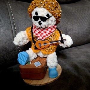 Può includere: Un orsacchiotto bianco fatto a mano con occhiali da sole, un cappello color oro e una sciarpa a scacchi rossi e bianchi. L'orso tiene in mano un piccolo ukulele ed è in piedi su una base di legno con una valigia marrone e stivali blu.
