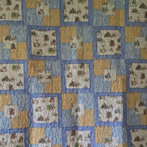 May include: A patchwork quilt with a repeating pattern of squares. The squares are in shades of blue, yellow, and white. The white squares feature a whimsical print of animals and figures. The quilt is likely handmade.