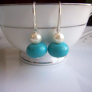 Op de afbeelding: Turquoise en witte hangende oorbellen met een zilveren haak. De turquoise kralen zijn rond en de witte kralen lijken op parels.
