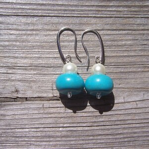 Op de afbeelding: Een paar turquoise en witte oorhangers met zilveren haakjes. De oorhangers hebben een ronde turquoise kraal en een kleinere witte kraal.