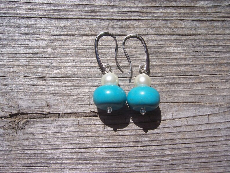 Op de afbeelding: Een paar turquoise en witte oorhangers met zilveren haakjes. De oorhangers hebben een ronde turquoise kraal en een kleinere witte kraal.