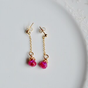 May include: A pair of gold dangle earrings with pink gemstone drops. The earrings feature a delicate gold chain and a small gold ball at the top.