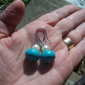 Op de afbeelding: Een paar zilveren oorhangers met turquoise en witte parels. De oorbellen zijn op een hand met een gouden ring.