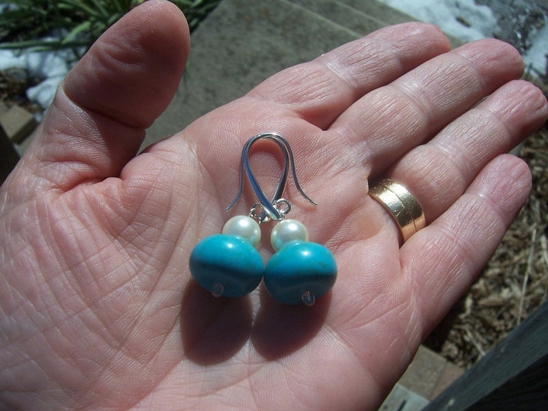 Op de afbeelding: Een paar zilveren oorhangers met turquoise en witte parels. De oorbellen zijn op een hand met een gouden ring.