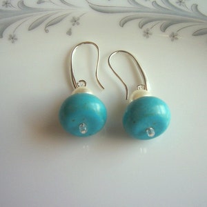 Op de afbeelding: Een paar zilveren oorhangers met turquoise en witte kralen. De turquoise kralen zijn rond en hebben een glanzende afwerking.
