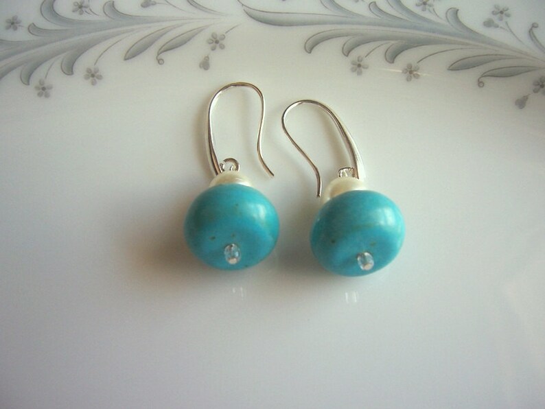 Op de afbeelding: Een paar zilveren oorhangers met turquoise en witte kralen. De turquoise kralen zijn rond en hebben een glanzende afwerking.