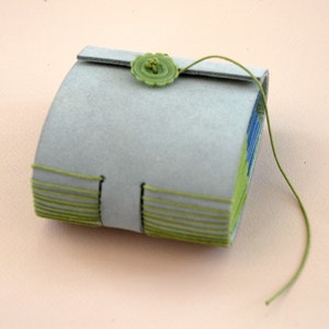 May include: A small, gray leather-bound journal with green stitching and a green button closure. The journal is closed and the stitching is visible on the spine.