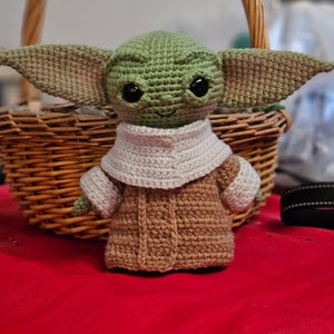 May include: A crocheted green and brown plush toy resembling a character from the Star Wars franchise. The toy has large ears, big eyes, and a small mouth. It is wearing a brown coat with a white collar.