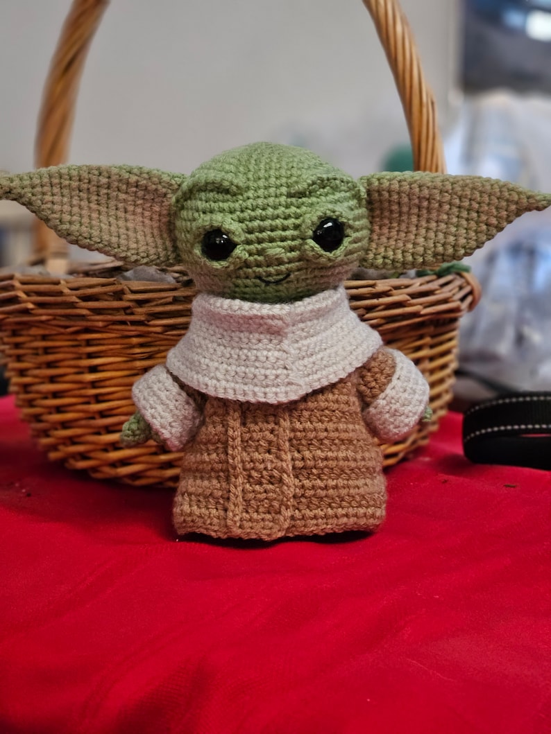 May include: A crocheted green and brown plush toy resembling a character from the Star Wars franchise. The toy has large ears, big eyes, and a small mouth. It is wearing a brown coat with a white collar.