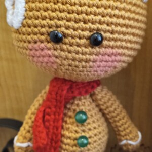 May include: A crocheted gingerbread man doll with a white icing top, red scarf, and green button accents.