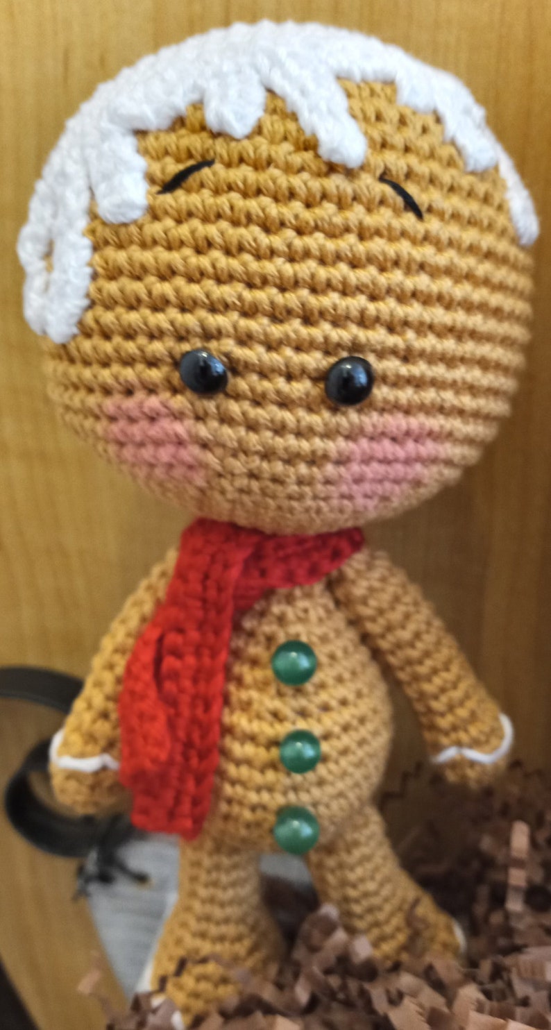 May include: A crocheted gingerbread man doll with a white icing top, red scarf, and green button accents.