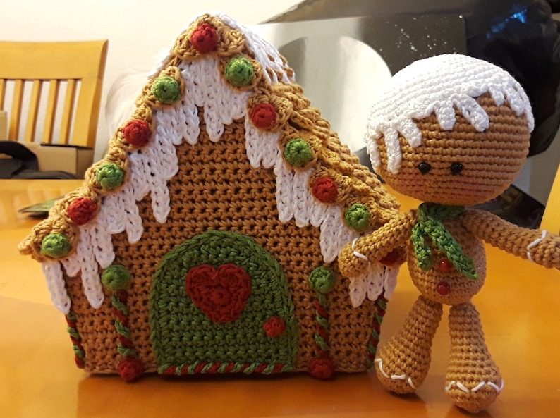 May include: A crocheted gingerbread house with white icing and red and green candy decorations. A crocheted gingerbread person stands next to the house.