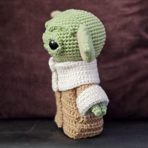 May include: A crocheted green and brown plush toy of a character from the Star Wars franchise. The toy is wearing a white crocheted coat.