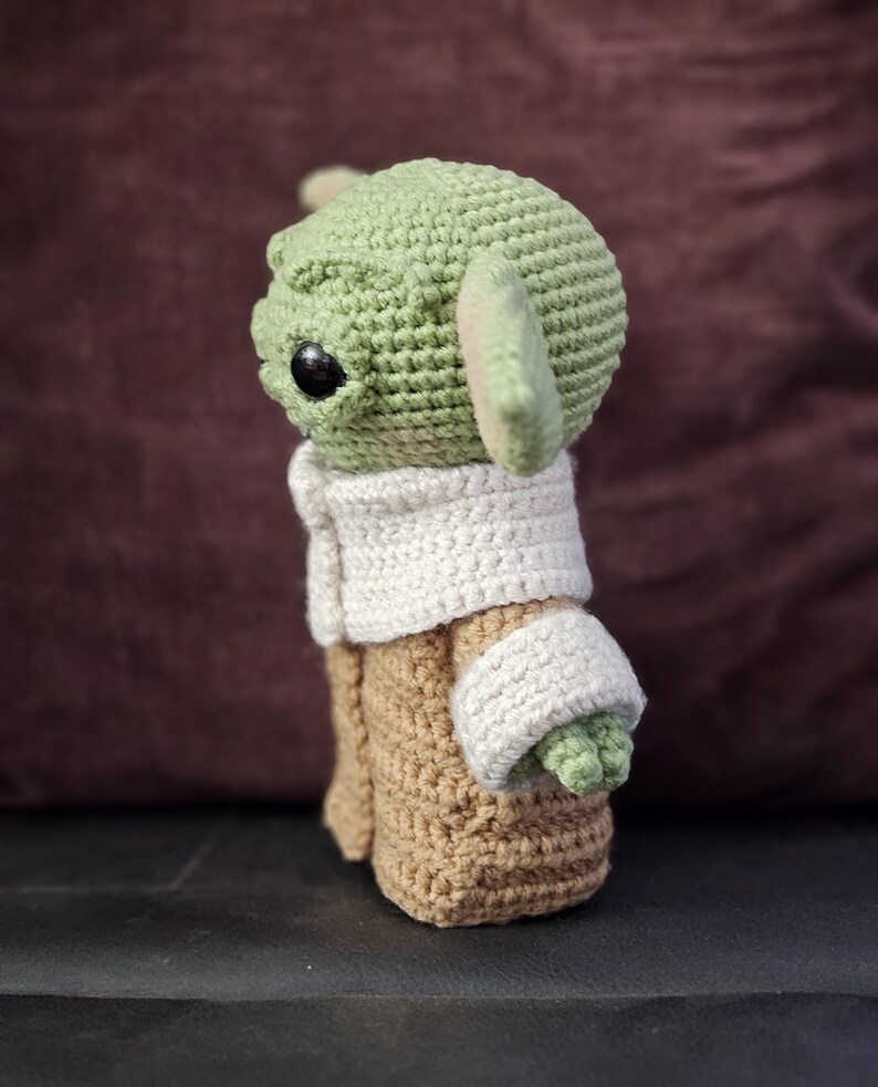 May include: A crocheted green and brown plush toy of a character from the Star Wars franchise. The toy is wearing a white crocheted coat.