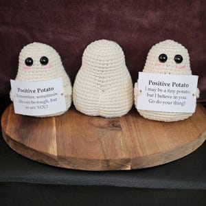 Puede incluir: Tres figuras de patata de ganchillo con ojos negros y sonrisas rosas están sobre una tabla de madera. Cada patata sostiene un pequeño letrero con mensajes positivos. El texto en los letreros dice "Positive Potato" y citas inspiradoras.