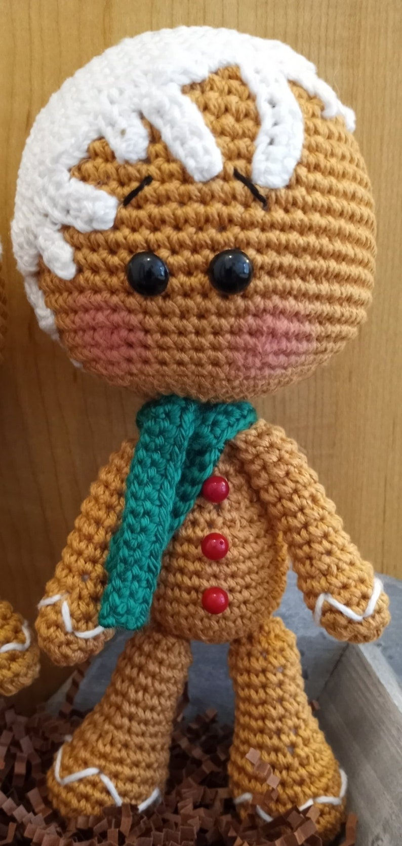 May include: A crocheted gingerbread man doll with white icing, green scarf, and red buttons. The doll is standing on brown shredded paper.