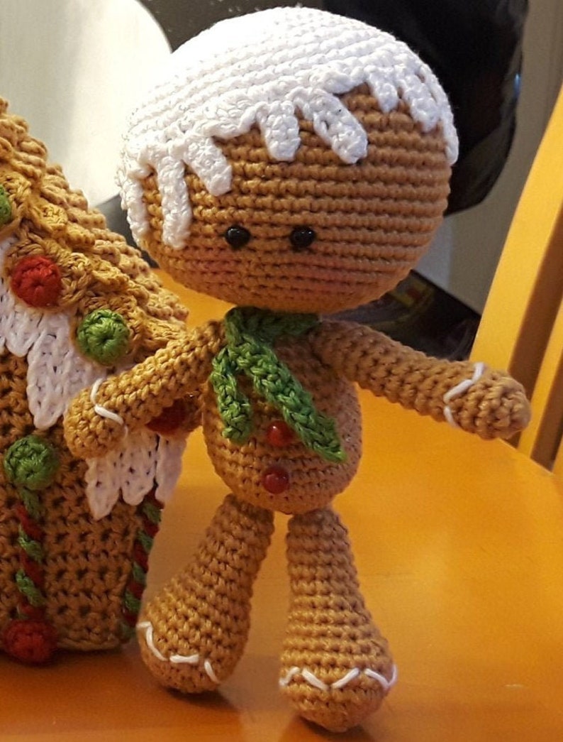 May include: A crocheted gingerbread man doll with white icing on its head, green scarf, and red buttons. The doll is standing next to a crocheted gingerbread house.