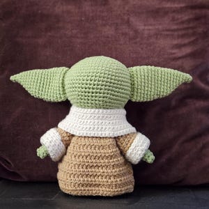 May include: A crocheted plush toy of a green character with large ears, wearing a brown and white crocheted outfit. The toy is sitting on a brown surface.