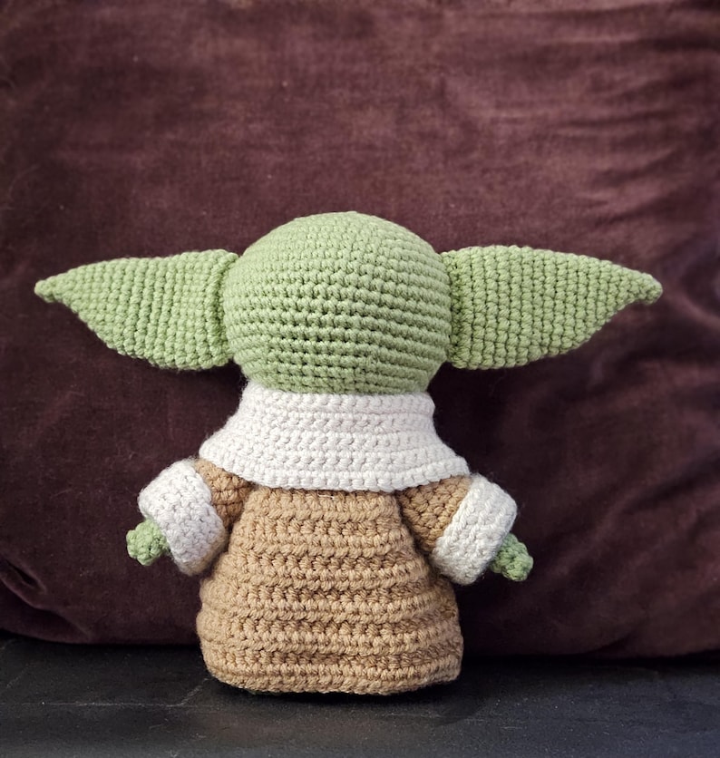 May include: A crocheted plush toy of a green character with large ears, wearing a brown and white crocheted outfit. The toy is sitting on a brown surface.