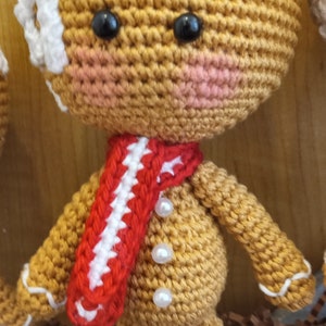 May include: A crocheted gingerbread man toy with white icing, red and white striped scarf, and pearl buttons. The toy is sitting on brown shredded paper.
