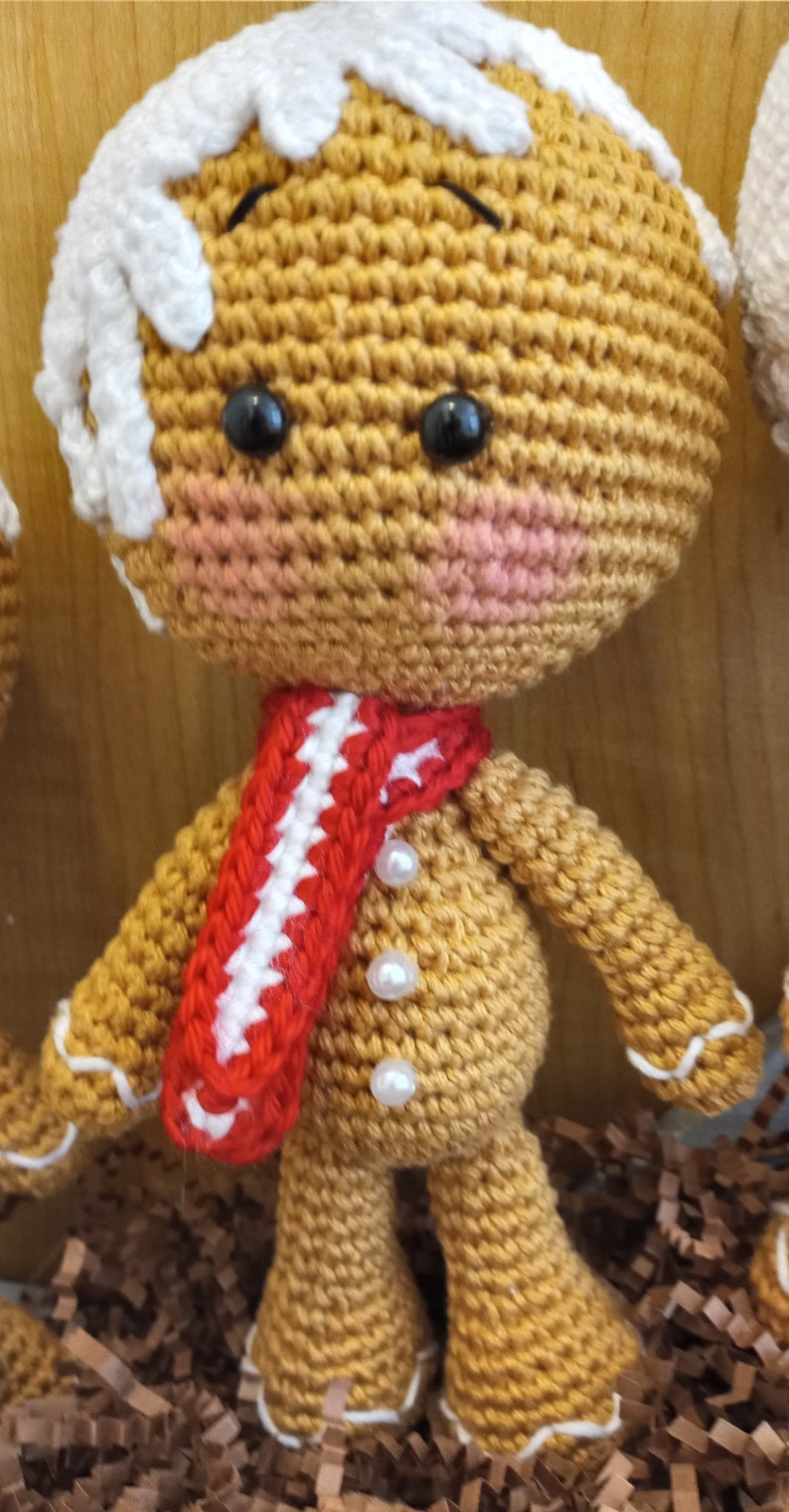 May include: A crocheted gingerbread man toy with white icing, red and white striped scarf, and pearl buttons. The toy is sitting on brown shredded paper.
