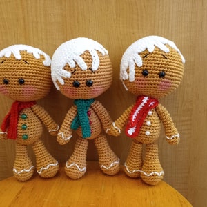 May include: Three crocheted gingerbread people toys. The gingerbread people are wearing white frosting hats and red, green, and white scarves.