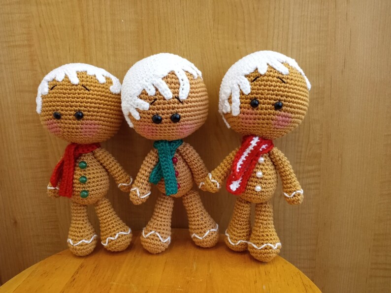 May include: Three crocheted gingerbread people toys. The gingerbread people are wearing white frosting hats and red, green, and white scarves.