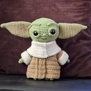 May include: A crocheted plush toy of a green alien character with large ears, wearing a brown coat with a white collar. The toy has big black eyes and a smiling mouth.
