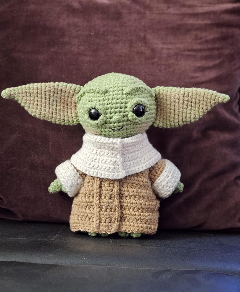 May include: A crocheted plush toy of a green alien character with large ears, wearing a brown coat with a white collar. The toy has big black eyes and a smiling mouth.