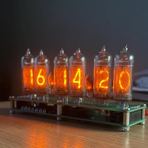 Könnte beinhalten: Eine Digitaluhr mit fünf Glasröhren, die die Uhrzeit 16:14:20 in leuchtend orangefarbenen Ziffern anzeigt. Die Uhr ist auf einer grünen Leiterplattenbasis montiert und steht auf einer Holzoberfläche.
