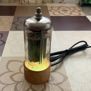 Könnte beinhalten: Eine Vintage-Nixie-Röhrenlampe mit einem transparenten Glasrohr und einem Holzsockel. Die Lampe hat eine silberne Oberseite und eine grüne Innenkomponente. Ein schwarzes Kabel erstreckt sich von der Basis. Die Lampe steht auf einer gemusterten Oberfläche.