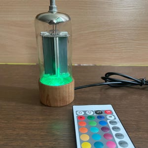 Könnte beinhalten: Eine dekorative Lampe mit einem Glasröhren-Design, mit einem Holzsockel und grünem Licht. Eine Fernbedienung mit verschiedenen Farboptionen und Einstellungen befindet sich neben der Lampe. Die Lampe hat eine silberne Oberseite und ein schwarzes Kabel.