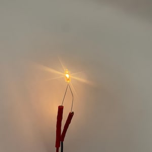 Könnte beinhalten: Eine kleine, klare Glühbirne, die ein helles, warmes orangefarbenes Licht ausstrahlt. Die Glühbirne ist mit zwei dünnen Metalldrähten verbunden, die an roten und schwarzen elektrischen Anschlüssen an der Basis der freiliegenden Verkabelung befestigt sind.