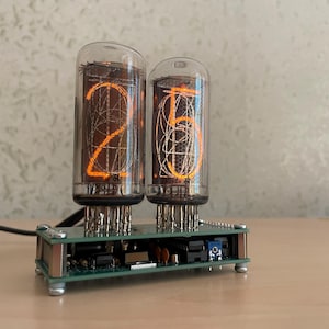 Könnte beinhalten: Eine Nixie-Röhrenuhr mit zwei vertikalen Glas-Vakuumröhren, die auf einer grünen Leiterplattenbasis montiert sind. Die Röhren leuchten in einem hellen Orange und zeigen die Zahl 25 mit sichtbaren internen Drahtfilamenten und Metallkomponenten.