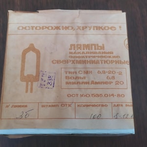 May include: A white cardboard box with a warning label that reads "ОСТОРОЖНО, ХРУПКОЕ!" in Cyrillic script. The box contains miniature light bulbs. The label also includes the text "ЛАМПЫ" and "СΒΕΡΧΜΗНИАТЮРНЫе" in Cyrillic script. The box has a label with the text "№ линии 36" and "количество 100".