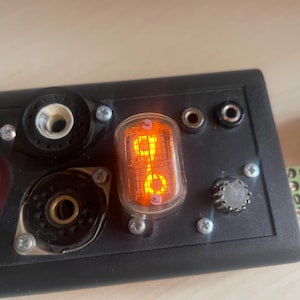 Peut inclure: Gros plan sur un panneau électronique noir avec diverses prises, vis et un tube Nixie orange éclairé affichant le chiffre 8. Le panneau a une esthétique vintage.